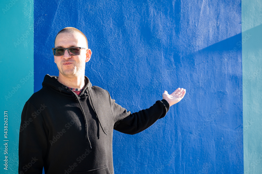 Stylish guy in glasses and black sweatshirt isolated on plain bright blue background, pointing with hand to side empty space for table or graph. Photo of model on wall background with empty space