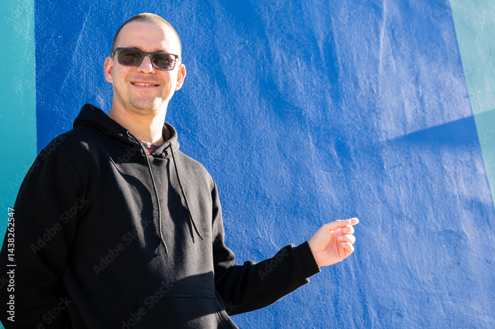 Stylish guy in glasses and black sweatshirt isolated on plain bright blue background, pointing with hand to side empty space for table or graph. Photo of model on wall background with empty space