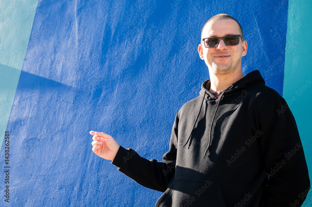Stylish guy in glasses and black sweatshirt isolated on plain bright blue background, pointing with hand to side empty space for table or graph. Photo of model on wall background with empty space