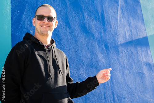 Obraz na płótnie Stylish guy in glasses and black sweatshirt isolated on plain bright blue background, pointing with hand to side empty space for table or graph. Photo of model on wall background with empty space w salonie