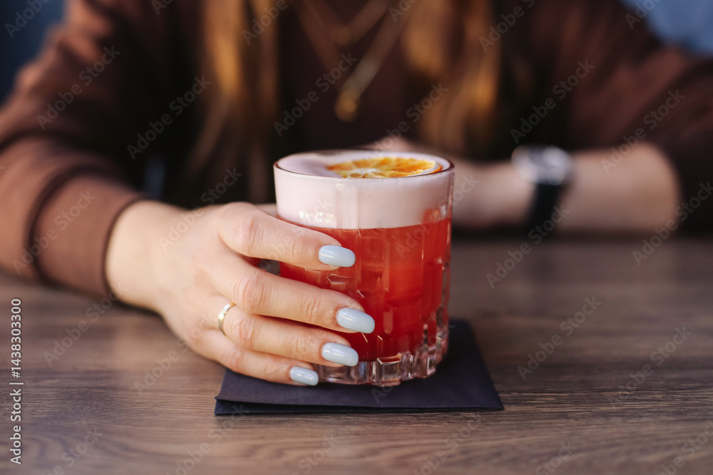 Refreshing red cocktail with foam. Elegant drink with citrus slice. Woman hand holding glass. Summer party drink. Fresh fruity cocktail. Modern bar atmosphere. Colorful evening beverage background.