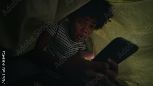 Medium shot of cheerful, excited 13-year-old African American boy lying in bed at night, hiding under duvet, viewing inappropriate or entertaining content on smartphone with surprise