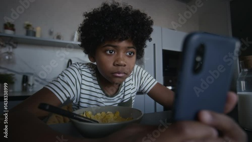 Medium shot of 13-year-old African American boy eating cornflakes in morning in kitchen, getting message with mature or inappropriate content, checking around, viewing it and grinning