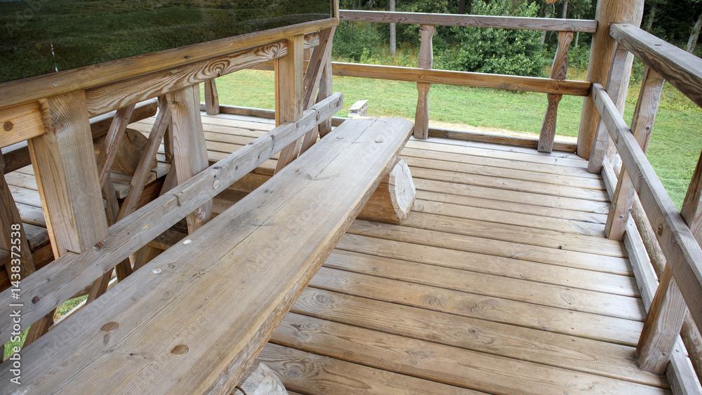 The interior of a wooden gazebo with a wooden floor, benches and railings, overlooking the green grass and trees outside.