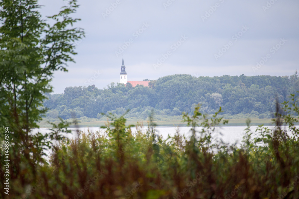 A landscape with a body of water in the foreground, behind which green trees spread out, and a building with a pointed tower, possibly a church, can be seen in the background.
