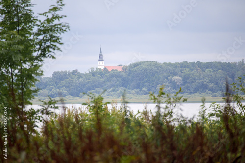 Obraz na płótnie
A landscape with a body of water in the foreground, behind which green trees spread out, and a building with a pointed tower, possibly a church, can be seen in the background. w salonie