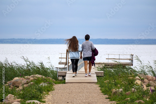 Obraz na płótnie
Two people, a man and a woman, walking from behind along a wooden pier towards a calm body of water under a cloudy sky. w salonie