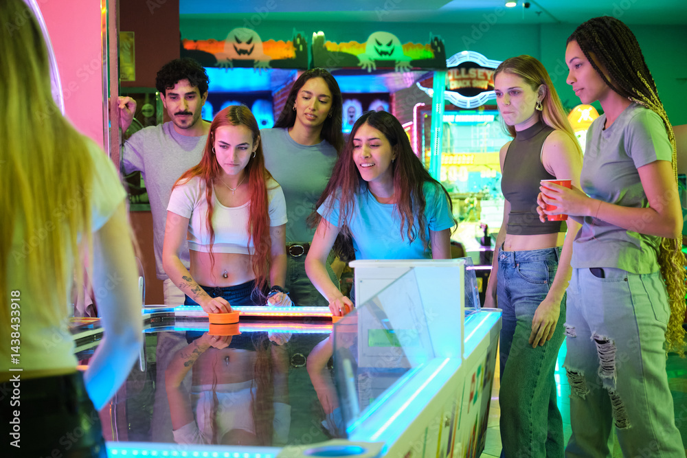 Young women competing on an air hockey table, surrounded by their friends, in a colorful amusement arcade