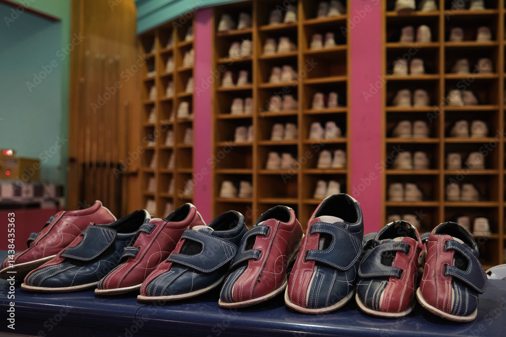 Several pairs of bowling shoes sitting on a counter, ready for bowlers, with a shoe rack filled with more shoes in the background