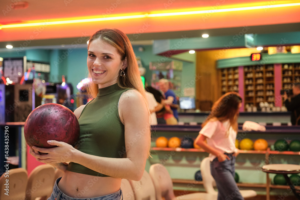 Happy young woman holding bowling ball, smiling in bowling alley, ready for a game with friends