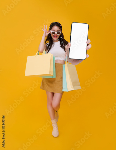 Happy beautiful Asian woman shopper showing smartphone mockup blank screen and hand carrying colorful shopping bags isolated on yellow background.