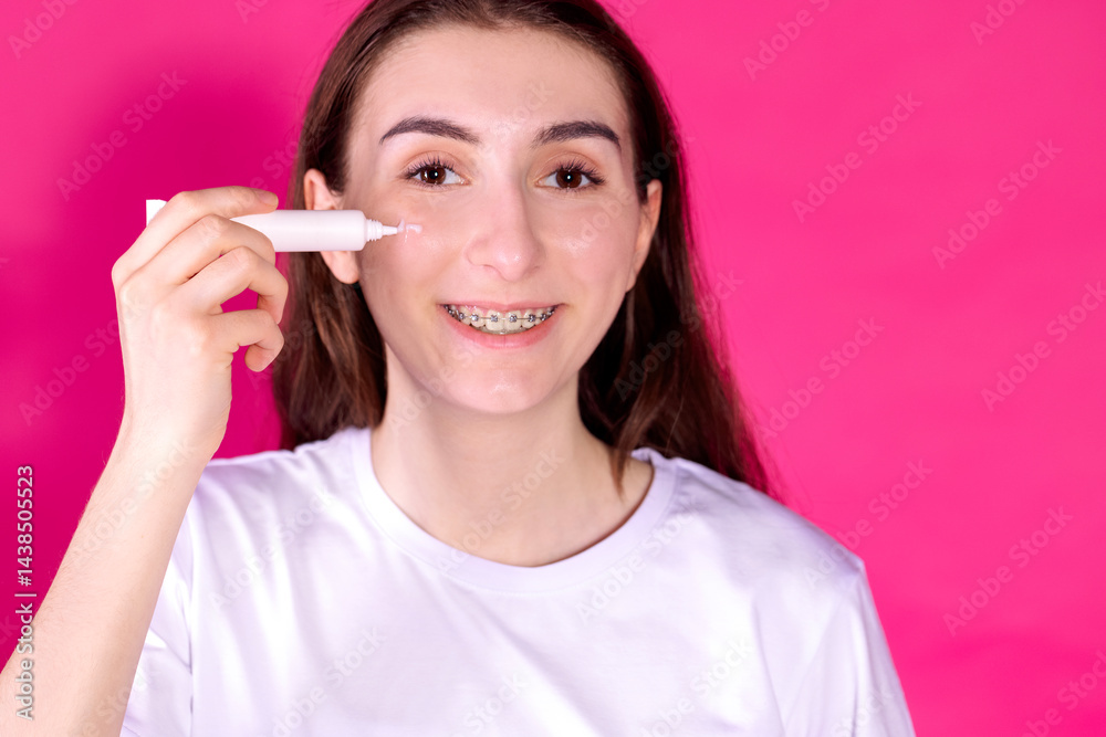 Close-up portrait of a young girl applying white cream on her face. The teenager smiles and holds a tube of cosmetics. Pink background. Skin care. Beauty treatments. Copy space. Close-up portrait of a young girl applying white cream on her face. The teenager smiles and holds a tube of cosmetics. Pink background. Skin care. Beauty treatments. Copy space.
