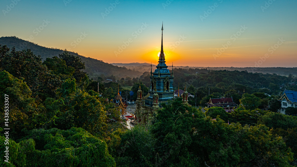 Fototapeta premium A breathtaking aerial view of a golden Buddhist temple at Wat Mae Klang Waterfall in Chiang Mai Thailand is surrounded by lush greenery, with the sunrise casting warm hues across the horizon.