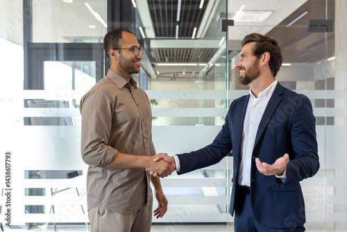 Two businessmen shake hands in a modern office, smiling warmly during a professional agreement or greeting, with natural light. photo