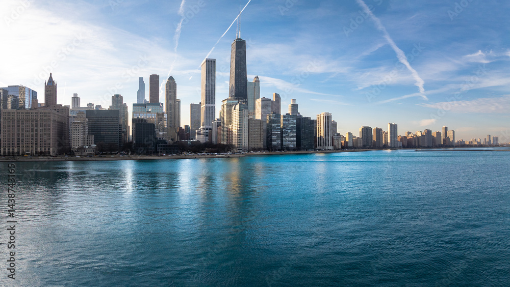 Fototapeta premium A stunning aerial view of the Chicago skyline along the Lake Michigan waterfront on a clear, sunny day. Skyscrapers rise above the shimmering blue water, with crisp contrails streaking across the vibr
