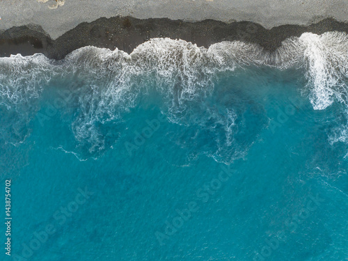 Waves gently lapping at a black sand beach under a clear blue sky at midday