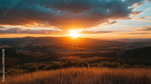 Sunset above landscape. Sun shining brightly. Sky with dark clouds. Golden light on grass and trees. Warm colors glow over the distant hills.