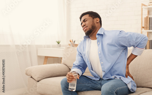 Young african-american man with back pain, pressing on hip with painful expression, sitting on sofa at home with glass of water, copy space