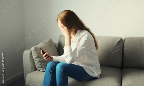 Sad upset crying woman holding phone suffering from stress after reading emotional message at home