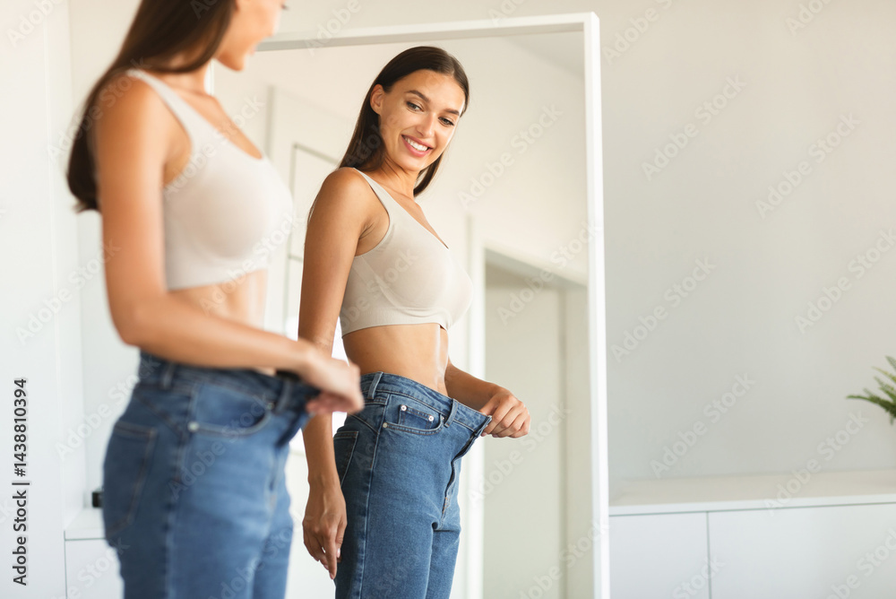 Fototapeta premium Excited young fit woman losing weight and wearing old too big jeans, woman feeling satisfied with results of her diet and slimming, posing near mirror, free space
