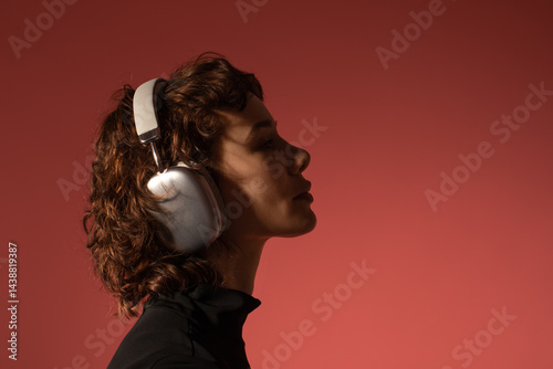Profile of young woman with curly hair wearing stylish headphones against red background. Relaxed mood, enjoying music, modern lifestyle and technology concept