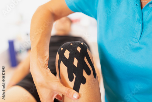 A female physiotherapist prepara the neuromuscular bandage or kinesiology tape from the knee of her patient, a young man, after an injury