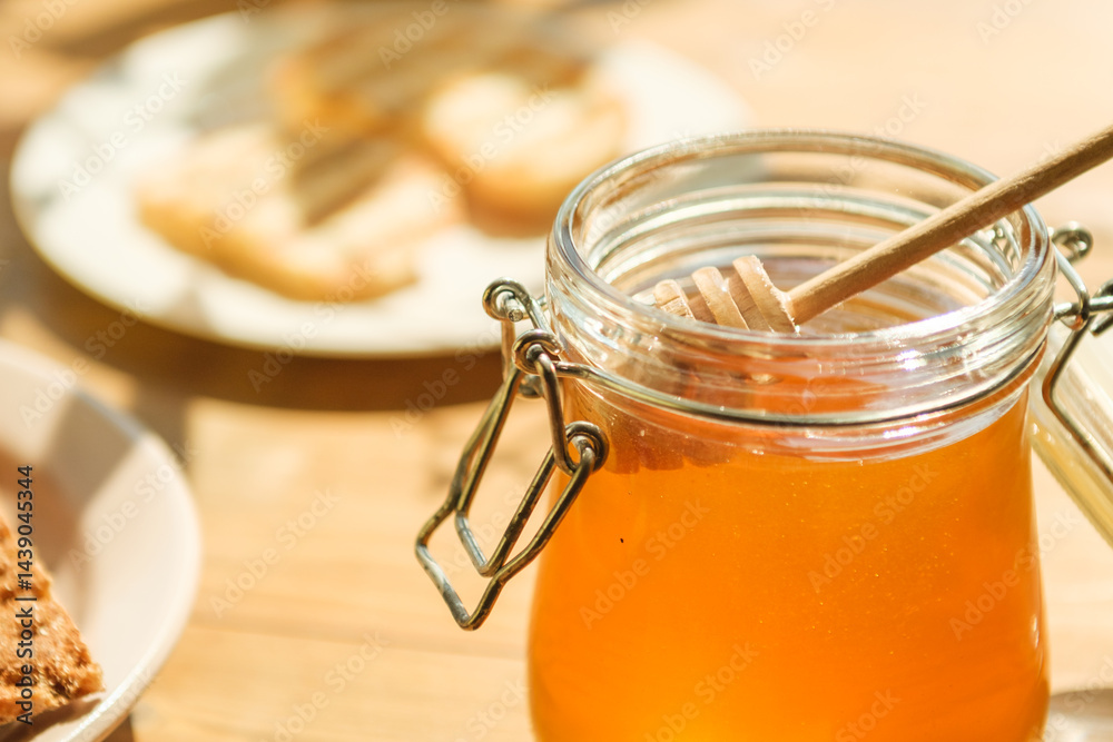 A jar of honey sits in a warm, sunlit kitchen with a wooden dipper resting inside. Next to it are slices of fresh bread on a plate, creating an inviting atmosphere.
