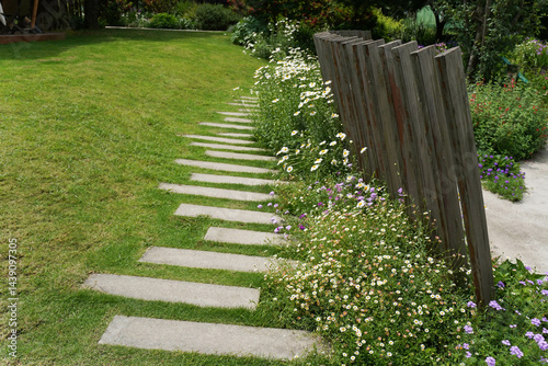 Garden path with stone slabs with green lawn along wooden fence