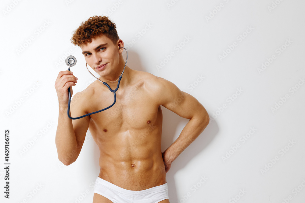 Fototapeta premium Confident young man in underwear holding a stethoscope, smiling while posing against a white background. Healthcare and fitness concept.