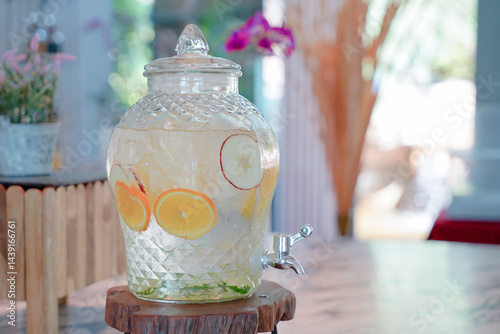 Fruit infused water in glass dispenser with citrus and apple slices, refreshing drink on wooden table in bright indoor setting