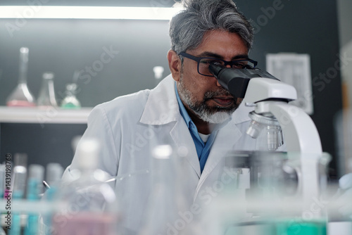 Middle-aged scientist examining samples under microscope in laboratory focusing on research. Various lab equipment and test tubes visible in background