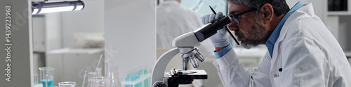 Scientist in laboratory using a microscope for in-depth analysis of samples. Laboratory equipment and beakers with blue liquids are visible in background