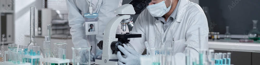 Researchers wearing lab coats and masks working with microscope and glassware in laboratory environment, focusing on scientific research and analysis