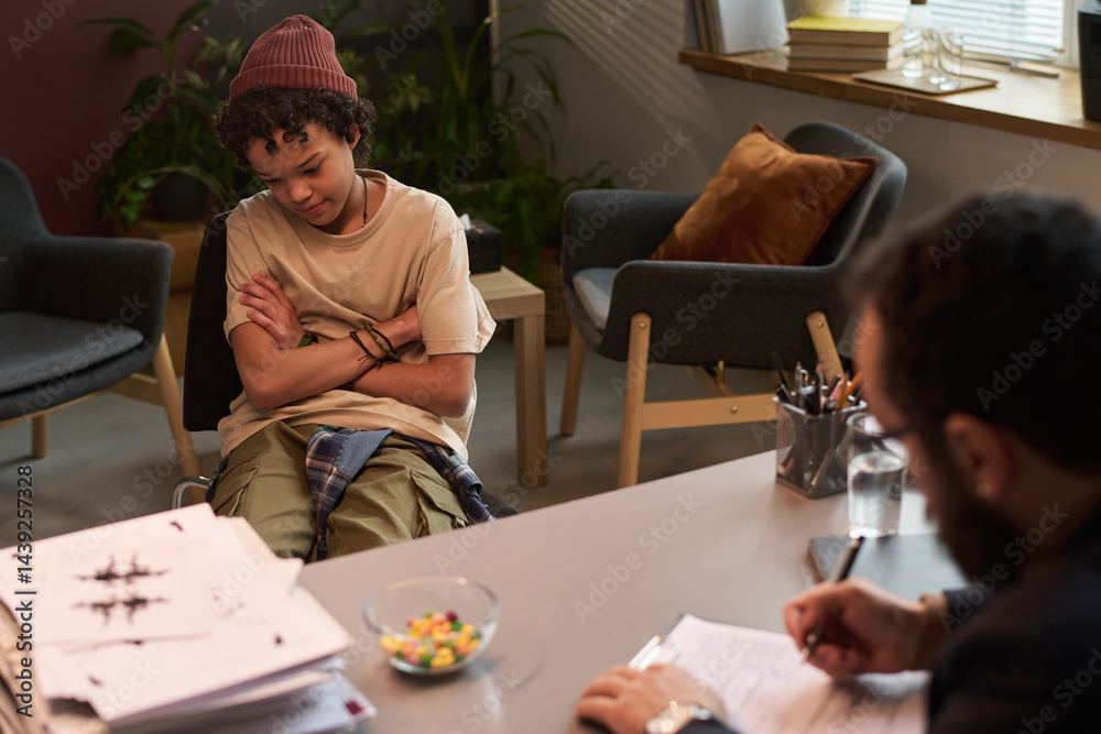 African American schoolboy crossing arms by chest and looking aside while sitting in front of school psychologist in office