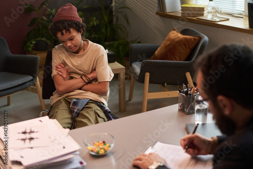 Obraz na płótnie African American schoolboy crossing arms by chest and looking aside while sitting in front of school psychologist in office w salonie