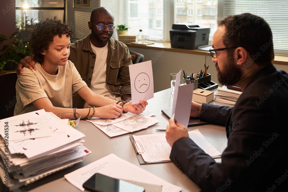 Cute schoolboy holding paper with sad smile while sitting in front of school psychologist during psychotherapy session