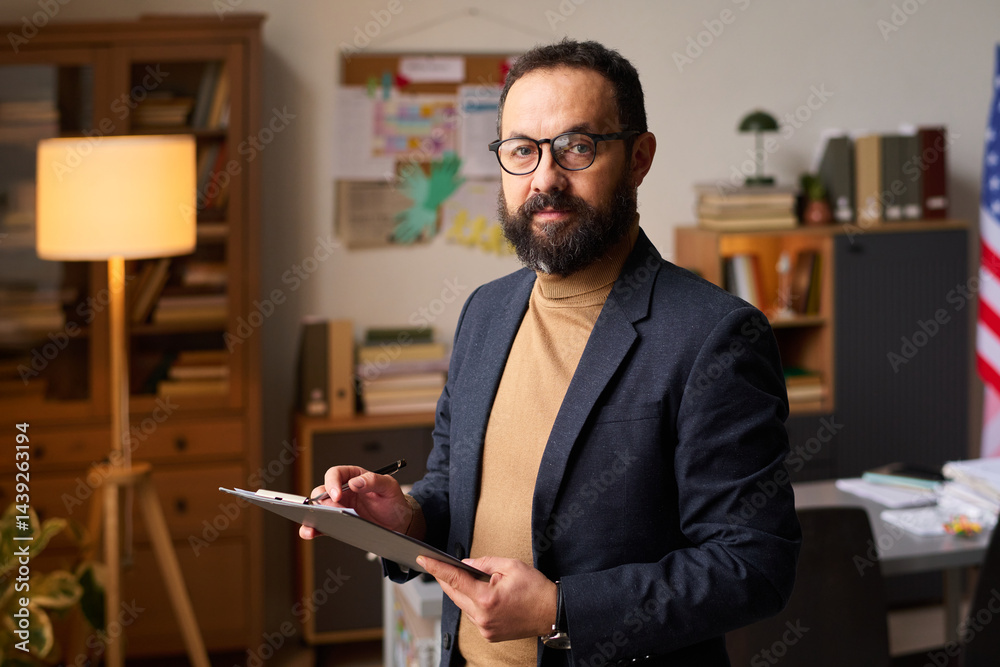Bearded male psychologist with clipboard looking at camera against his workplace while working in office at school