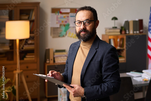 Obraz na płótnie Bearded male psychologist with clipboard looking at camera against his workplace while working in office at school w salonie