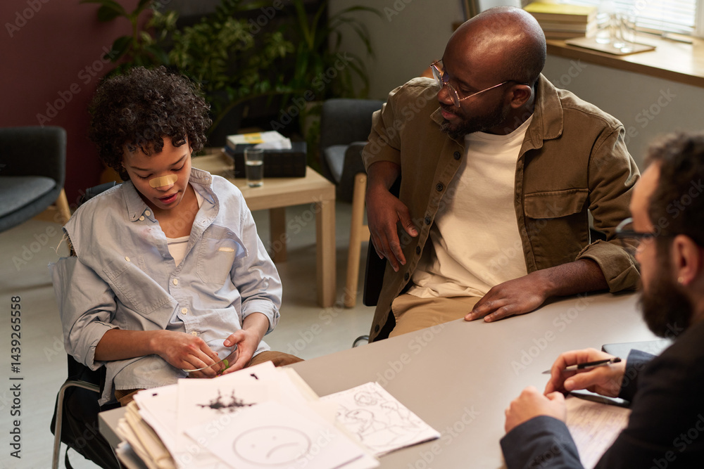 Naughty African American boy sitting next to his father during discussion of his fight with classmates at psychological session