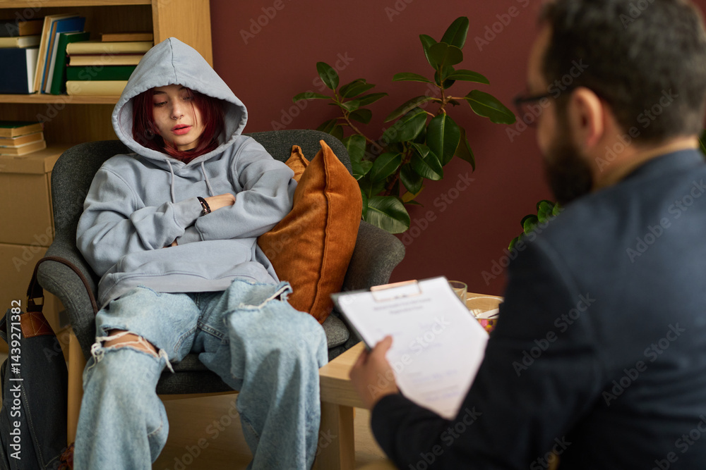 Youthful girl in casual attire describing her problem to school psychologist with document sitting in front of patient