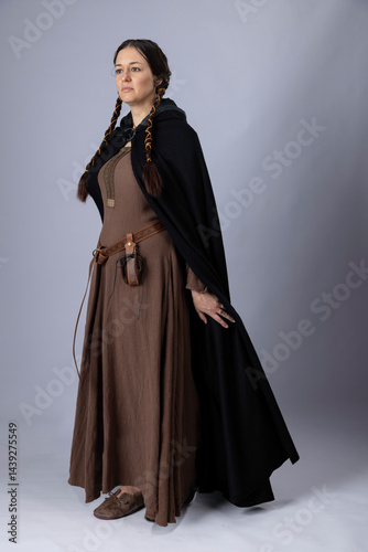 A Medieval peasant woman wearing a rough linen dress called a kirtle with a leather belt and pouch. She has long, dark hair arranged into long plaits
