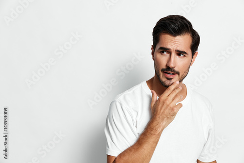 Thoughtful young man in white t-shirt touching his beard, pondering, looking sideways with a curious expression against a plain solid background. Lifestyle concept.