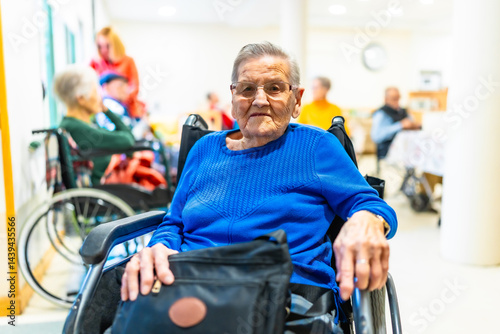 Elderly woman sitting in wheelchair in nursing home with other seniors in background