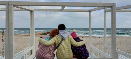 Three people in warm jackets and hats sitting closely on a white wooden bench, embracing and gazing at the waves of a cloudy sea, creating a cozy and emotional winter beach scene