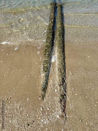 Two barnacle-covered pipes extend from sandy beach into clear shallow water, with small foamy waves under bright sunlight. From up