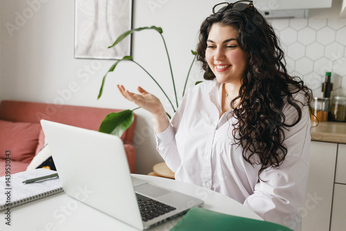 Side view of young caucasian female tutor having group video chat, explaining topic or new subject, gesticulating sitting at table in front of laptop at kitchen. Remote education concept