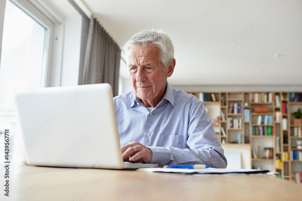Senior man working on a laptop at home with documents and bookshelf in the background Senior man working on a laptop at home with documents and bookshelf in the background