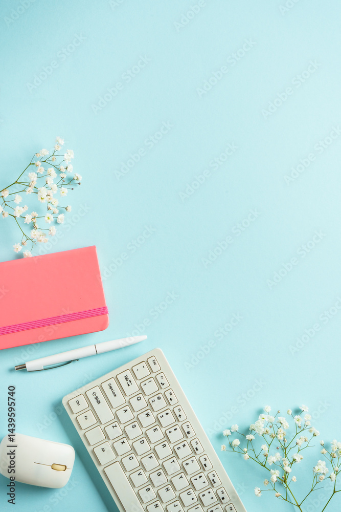Office desk with keyboard, notebook, pen and flowers on blue background. Flat lay with copy space.