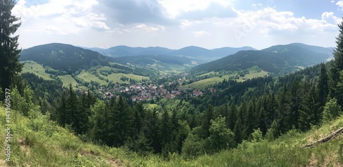 Panoramic view of a valley town nestled amongst lush green mountains and forests under a partly cloudy sky