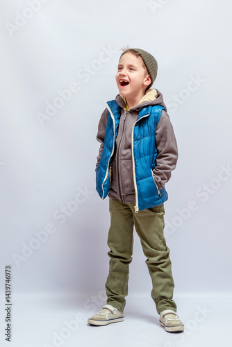 Young boy laughing, wearing a green beanie, blue vest, gray hoodie, and olive pants, standing casually with hands in pockets. Light-colored shoes. Joyful, cheerful expression